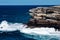 Oceanside rocky sandstone cliff with blue sea water waves creating whitewash against coastline and clear sky in background