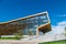 Office building in the meadow with glass windows that reflect the floating clouds, along the building there are people