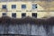 An old abandoned building with broken windows behind a fence of overgrown plants