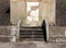 Old abandoned concrete shelter with a blocked door with cracked repaired steps and patched eroded walls in sunlight and shadow