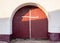 Old ancient entrance maroon gate door with a white cross on them against a solid white brick wall. Entrance to the Christian monas