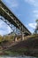 Old arch bridge viewed from beneath showing severe deterioration through rusting structural supports and crumbling concrete piers