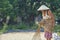 An old Asian woman wearing a hat is drying the harvested rice seeds in the hot sun