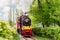 Old bright train in green city park on touristic railroad. Retro locomotive with clouds of steam from smoke pipe in forest