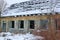 Old brown ruined brick house with empty black windows in white snow overgrown with plants and trees