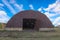 Old brown rusty metal hangar with open broken gates under a bright blue sky with white clouds