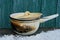 An old burnt saucepan covered by a lid stands on a wooden table in white snow against a green wall in the street