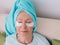 Old caucasian stylish woman laying on a couch with blue towel on her had and collagen eye patches.