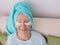 Old caucasian stylish woman laying on a couch with blue towel on her had and collagen eye patches.