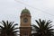 an old copper topped clock tower against an overcast white sky flanked by tropical palm trees along the seaside promenade in