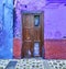 Old damaged wooden door and vibrant purple wall. Vintage weathered and damaged entrance to a colorful house in a small