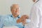 An old fat asian gray hair patient wearing glasses and light blue shirt sitting on hospital bed receive a glass of water from