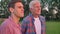 Old father hugging adult son and looking forward, standing on wheat or rye field, forest in background