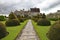 An old flag stone path leads up to an English country house. The path is bordered by elegant topiary bushes