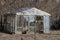 An old gray glass greenhouse stands outside in dry vegetation