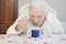 An old gray-haired woman sits at a table and drinks tea from a teaspoon inclined over a mug.