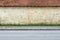 Old grunge yellow brick wall with terracotta tiled roof. A stripe of grass, concrete tiled sidewalk and asphalt road in front.