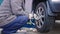 old man in dirty blue pants and grey jacket removing tire screws out of silver car