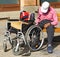 Old man sits on a bench next to a wheelchair