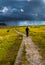 Old Man With Walking Stick Strolls On Narrow Gravel Path Through Scenic Landscape With Spectacular Clouds