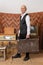 Old man in white shirt holds old classic travel trunk luggage, stands among vintage room with old tv and radio