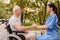 An old man who sits in a wheelchair and a nurse who sits next to the bench holding hands