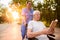 An old man who sits in a wheelchair and a nurse who is standing behind him reading a book in the park at sunset