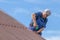 Old man working at heat on a roof of a house with electric screwdriver, wearing no safety devices, work clothing, blue overall, da