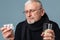 Old pensive sick man with gray hair and beard in eyeglasses and scarf thoughtfully looking on pills holding glass of