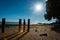 Old pier posts in silhouette cast shadow on sand at low tide with lens flare from starburst sun