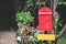 Old red wooden mailbox on black pillar with blank yellow wooden sign in the garden background, copy space