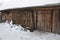 An old rustic rural wooden barn with a roof covered with snow and fire wood stacked along the barn wall in winter. Well-kept