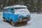 An old rusty ambulance van stands in a snow-covered clearing on a cloudy winter day.