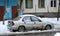 An old rusty gray car in snow and melt water is parked in the courtyard of a residential building