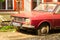 Old, rusty, red Dacia 1300 automobile, with a flat tire but active license plates, parked in the sun