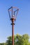 Old rusty street lamp with broken bulb and torn electrical wires against bright, summer blue sky and green trees. Devastation and