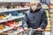 Old senior european man wearing protective facial mask pushing shopping cart in the supermarket. Shopping during COVID-19 concept