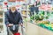 Old senior european man wearing protective facial mask pushing shopping cart in the supermarket. Shopping during COVID-19 concept