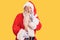 Old senior man with grey hair and long beard wearing traditional santa claus costume hand on mouth telling secret rumor,