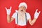 Old senior man with grey hair and long beard wearing white t-shirt and santa claus costume shouting with crazy expression doing