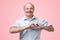 Old senior man with mustache making out of hands heart over pink background.