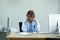 Old short blonde hair woman doctor with light blue shirt and white coat sitting on black chair in bright office in front of