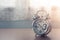 Old silver alarm clock on wooden table in bedroom with white curtain in the background.