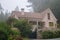 Old stone house with beautiful garden in morning fog. Rural cottage in village in forest. Cozy residence with trees and flowers.