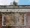 Old stone poster frame on the wall of a derelict cinema with plants growing out of a decaying brick wall and a blue cloudy sky