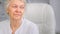 Old wise grey haired mother with blue eyes looks aside with sad face sitting in white chair under bright light