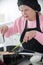 An old woman in a cap and apron frying pancakes and turning it over using a silicone paddle