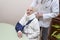 Old woman in a white bathrobe sits on a chair with a sling on her hand. The doctor puts on her neck an orthopedic collar.