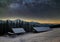 Old wooden house, hut and barn, pile of firewood in deep snow on mountain valley, spruce forest, woody hills on dark starry sky