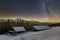 Old wooden house, hut and barn, pile of firewood in deep snow on mountain valley, spruce forest, woody hills on dark starry sky